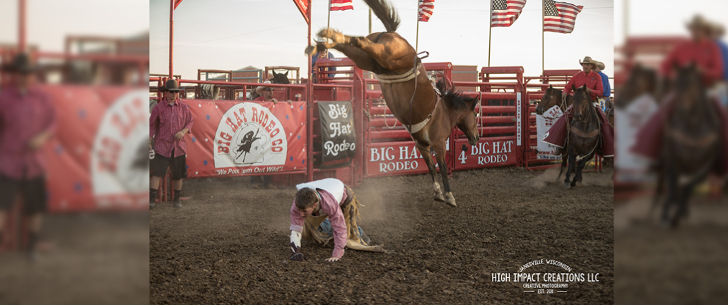 Bronc rider picks himself up from the arena floor as a bronc bucks in the background.